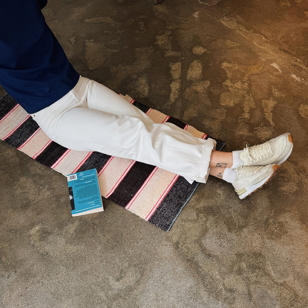 Person wearing white sneakers and light-colored pants relaxing on a handmade striped rag rug with a book next to them.