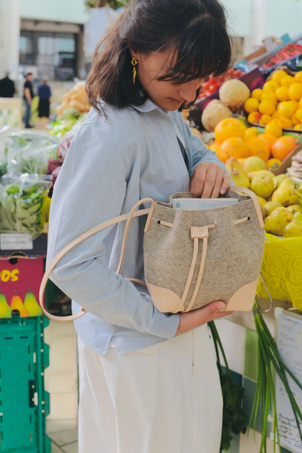 Woman holding a beige wool felt bucket bag in a market setting with fruits and vegetables.