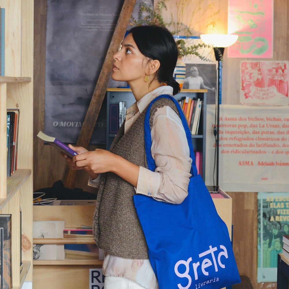Samarra vest. Woman wearing a knitted wool vest holding a book and a blue 'Greta' bag in a bookstore.