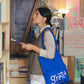 Samarra vest. Woman wearing a knitted wool vest holding a book and a blue 'Greta' bag in a bookstore.