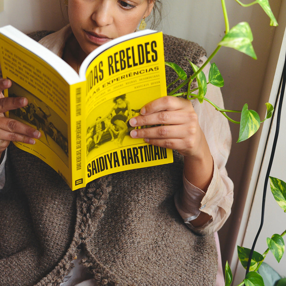 Samarra vest. Person wearing a knitted wool vest and reading a book titled 'Rebeldes' by Saidiya Hartman, surrounded by green plants.