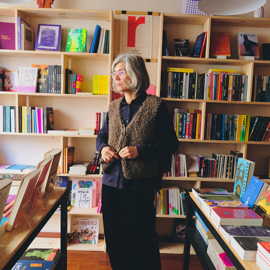 Samarra vest. Woman wearing a knitted wool vest standing in a bookstore surrounded by bookshelves filled with books.