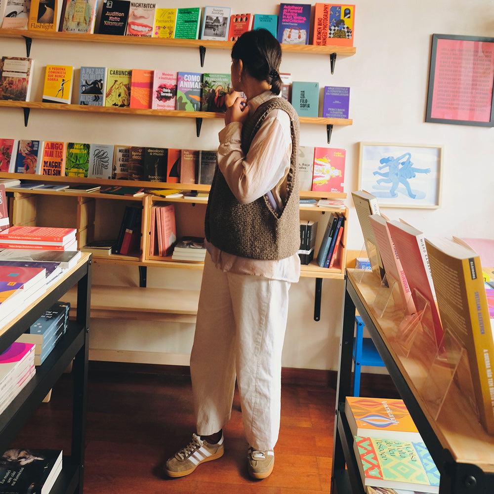 Person wearing a knitted wool vest and browsing books in a bookstore with shelves filled with colorful books.
